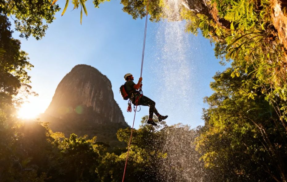 Um aventureiro pratica rapel em uma cachoeira na Mata Atlântica. Ele está suspenso por cordas, com a água caindo ao seu lado e a silhueta da Pedra do Bispo ao fundo, iluminada pelo sol poente.