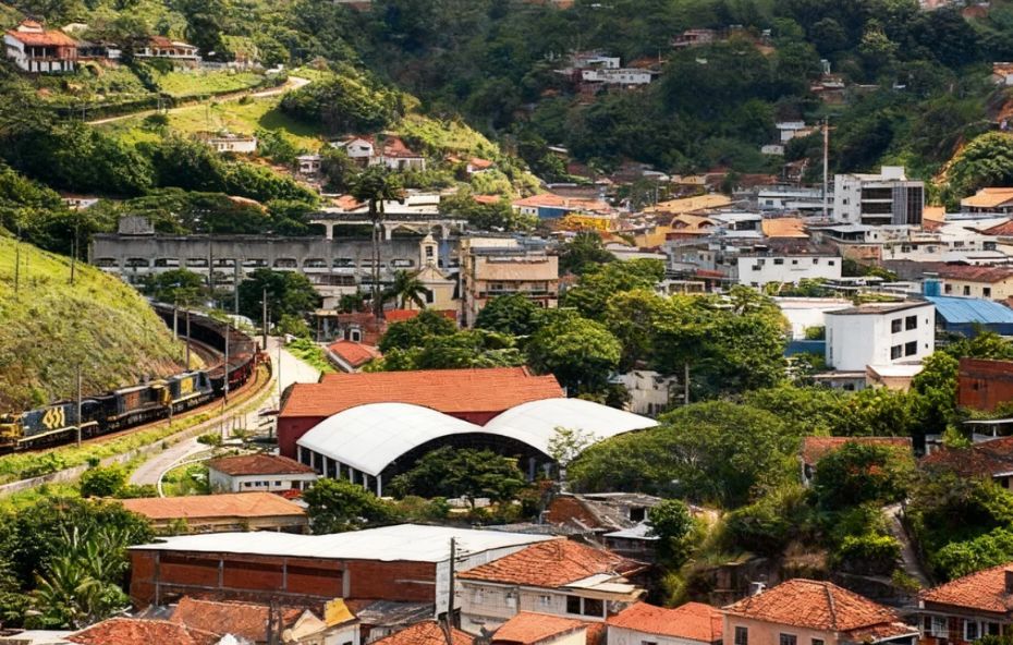 Vista panorâmica da cidade de Mendes, com suas casas e construções cercadas por montanhas verdes e uma linha de trem contornando a paisagem.