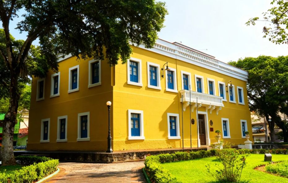 Fachada do Paço Municipal de Piraí, um casarão histórico de dois andares com pintura amarela e janelas azuis, rodeado por um jardim verde e árvores.