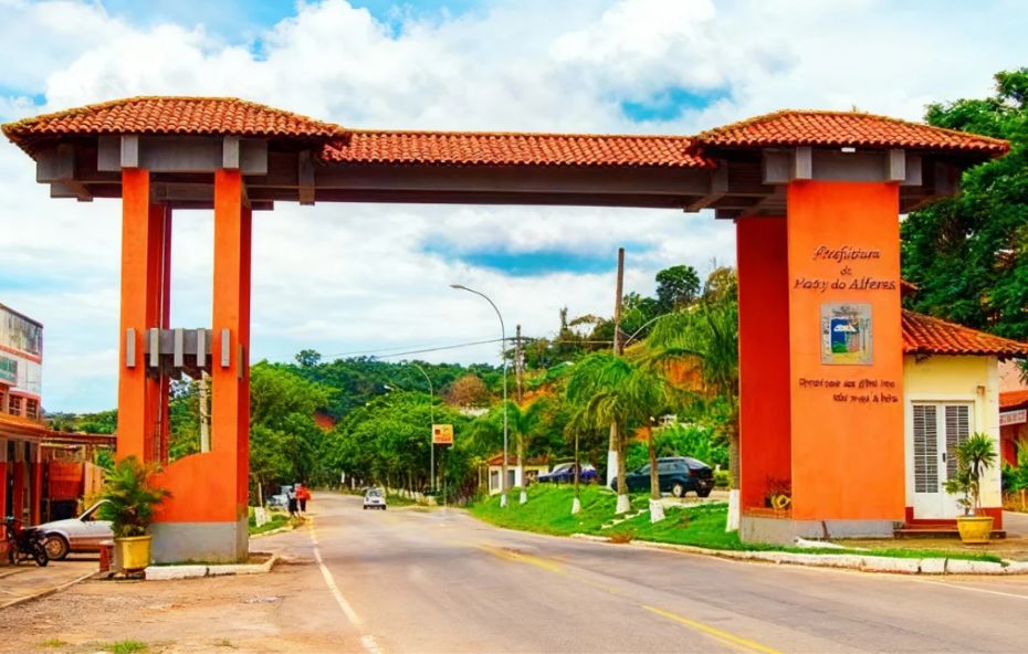 Imagem do pórtico de entrada de Paty do Alferes. A estrutura é laranja com telhado de estilo colonial. Ao fundo, morros verdes e céu azul com nuvens brancas.