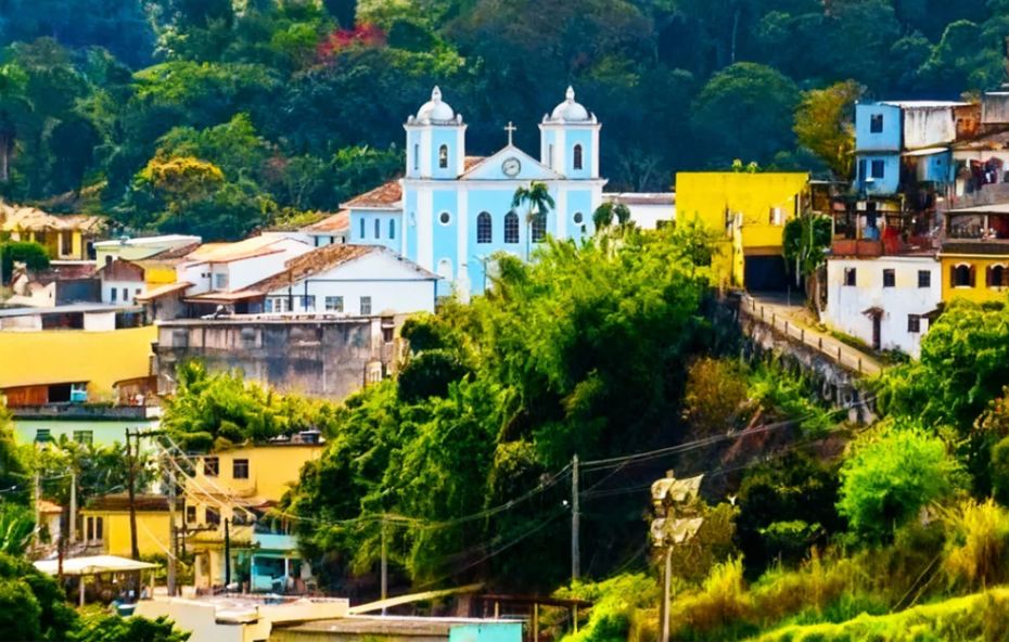Vista panorâmica da cidade de Rio Claro, com a igreja azul de São João Batista ao centro, cercada por casas coloridas e pela vegetação exuberante das montanhas.