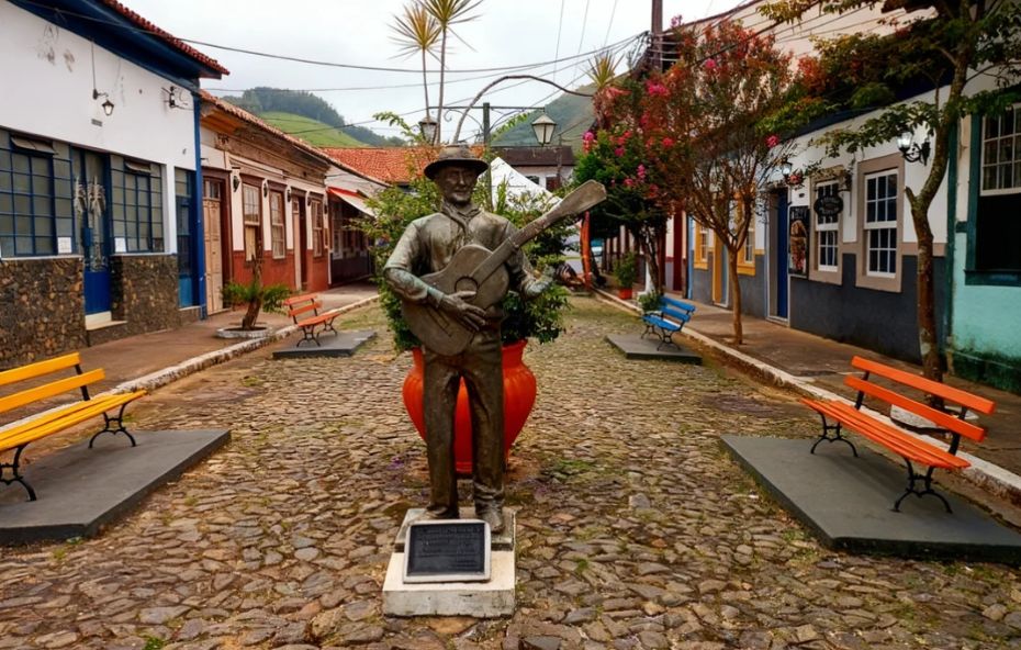 Imagem da rua principal de Conservatória. No centro, uma estátua de bronze de um violeiro. A rua é de paralelepípedos e ladeada por casarões coloniais coloridos e bancos de praça. Ao fundo, veem se morros e vegetação.