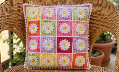 Almofada quadrada de crochê feita com quadradinhos coloridos e aplicação de margaridas brancas em relevo apoiada em cadeira de vime.