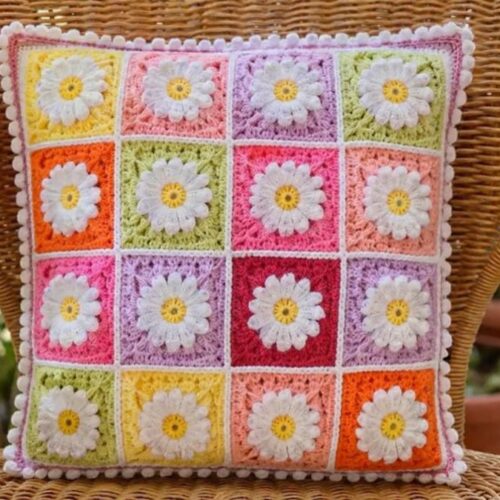 Almofada quadrada de crochê feita com quadradinhos coloridos e aplicação de margaridas brancas em relevo apoiada em cadeira de vime.