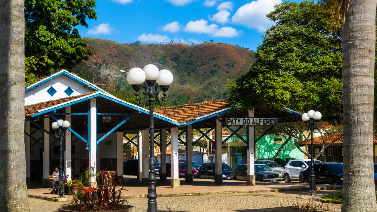 Fotografia da antiga estação de trem de Paty do Alferes, mostrando a estrutura de telhado colonial com o nome da cidade, postes de iluminação antigos, carros estacionados e montanhas ao fundo.