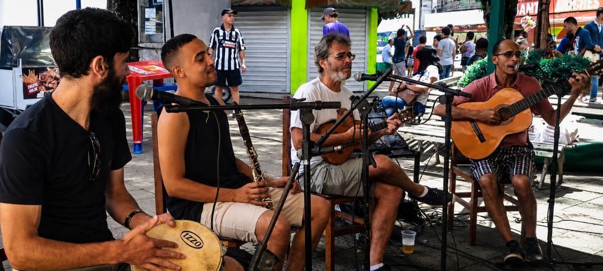 Quatro músicos tocam instrumentos como pandeiro, clarinete, cavaquinho e violão ao ar livre na Praça Doutor João Neri em Mendes.