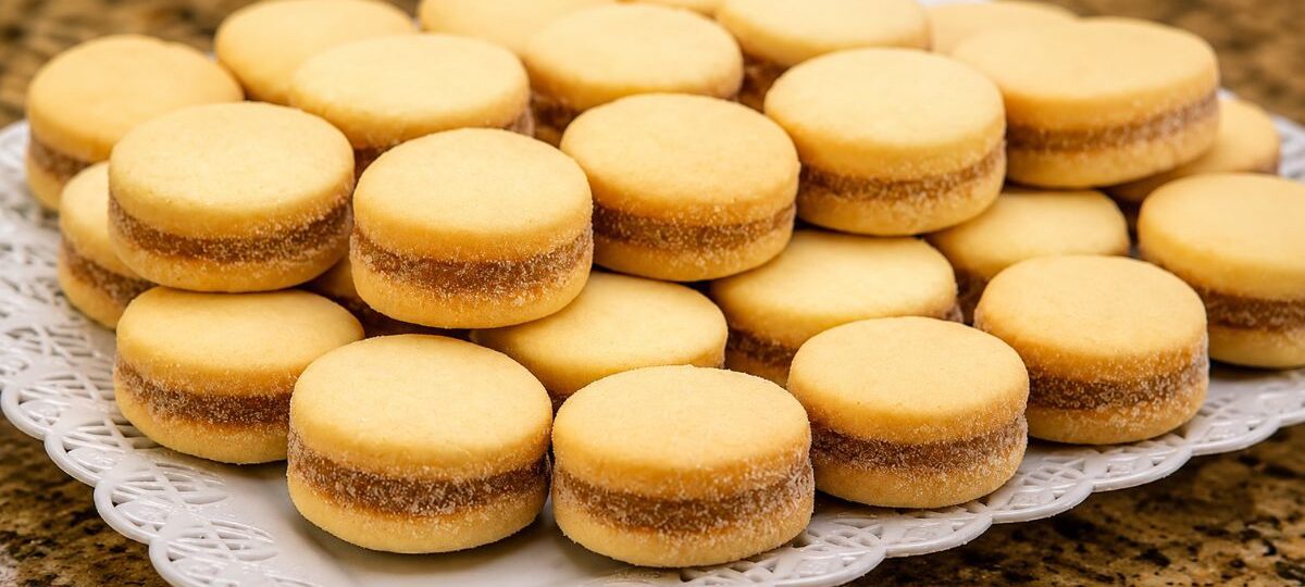 Biscoitos caseiros da roça redondos e dourados recheados com goiabada dispostos em uma travessa branca rendada.