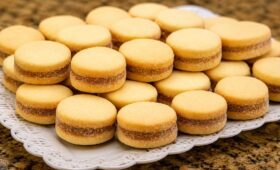 Biscoitos caseiros da roça redondos e dourados recheados com goiabada dispostos em uma travessa branca rendada.