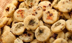 Biscoitos salgados caseiros amanteigados em formato redondo, polvilhados com ervas e pimenta em flocos, dispostos em uma cesta.