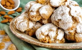 Biscoitos caseiros de amêndoas com cobertura rústica de açúcar em pó polvilhado, dispostos em um prato de cerâmica sobre pano verde com amêndoas ao fundo.