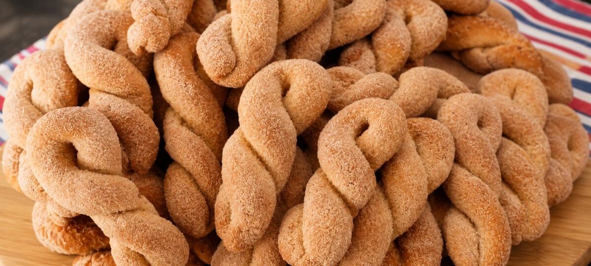 Uma pilha generosa de biscoitos de banana caseiros trançados, dourados e polvilhados com uma mistura de açúcar e canela, dispostos sobre uma tábua de madeira redonda.