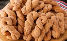 Uma pilha generosa de biscoitos de banana caseiros trançados, dourados e polvilhados com uma mistura de açúcar e canela, dispostos sobre uma tábua de madeira redonda.