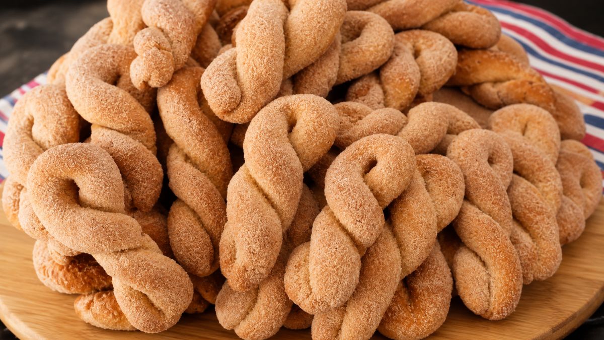 Uma pilha generosa de biscoitos de banana caseiros trançados, dourados e polvilhados com uma mistura de açúcar e canela, dispostos sobre uma tábua de madeira redonda.
