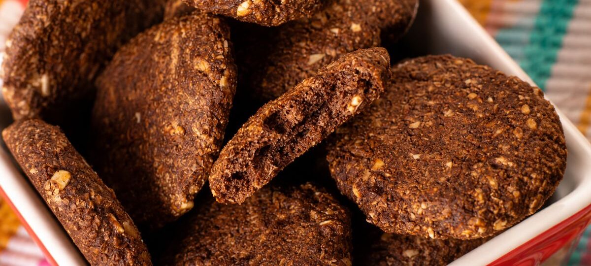 Pilha de biscoitos de cacau artesanais e crocantes com pedaços visíveis de castanhas em uma tigela vermelha sobre tecido colorido.