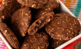 Pilha de biscoitos de cacau artesanais e crocantes com pedaços visíveis de castanhas em uma tigela vermelha sobre tecido colorido.