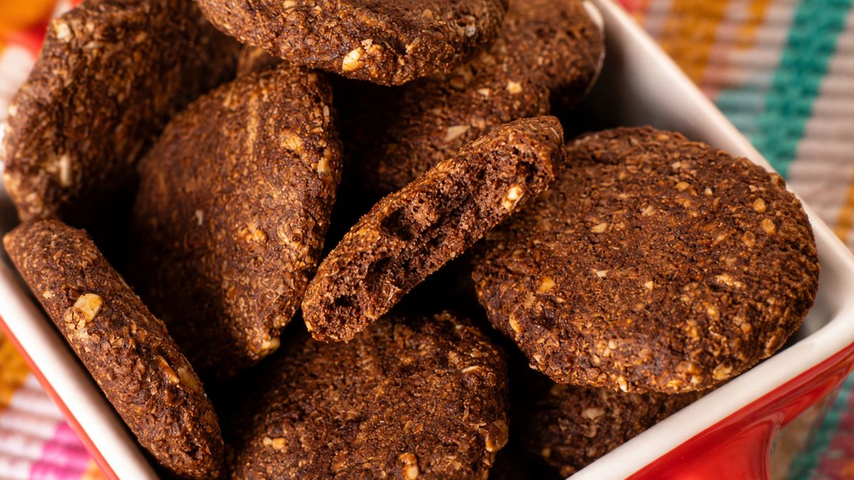 Pilha de biscoitos de cacau artesanais e crocantes com pedaços visíveis de castanhas em uma tigela vermelha sobre tecido colorido.