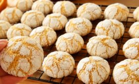 Uma fileira de biscoitos de laranja artesanais dourados e craquelados com açúcar de confeiteiro, dispostos sobre uma grade de resfriamento de metal preta em uma mesa de madeira. Uma mão segura um biscoito em primeiro plano.