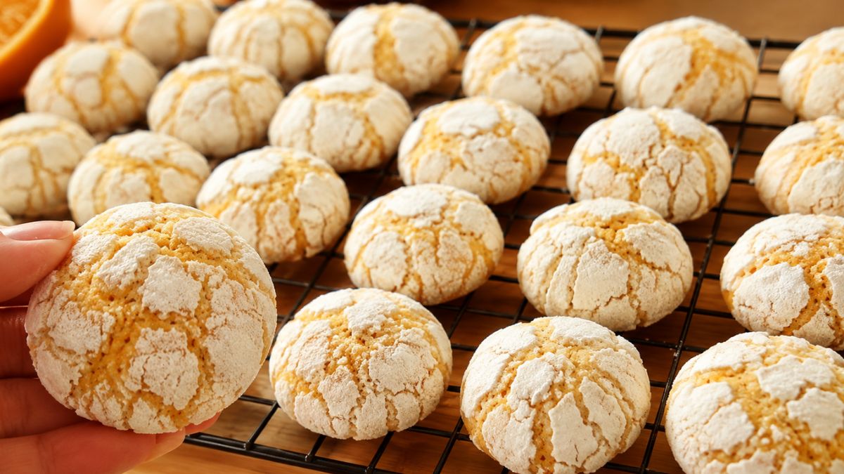 Uma fileira de biscoitos de laranja artesanais dourados e craquelados com açúcar de confeiteiro, dispostos sobre uma grade de resfriamento de metal preta em uma mesa de madeira. Uma mão segura um biscoito em primeiro plano.
