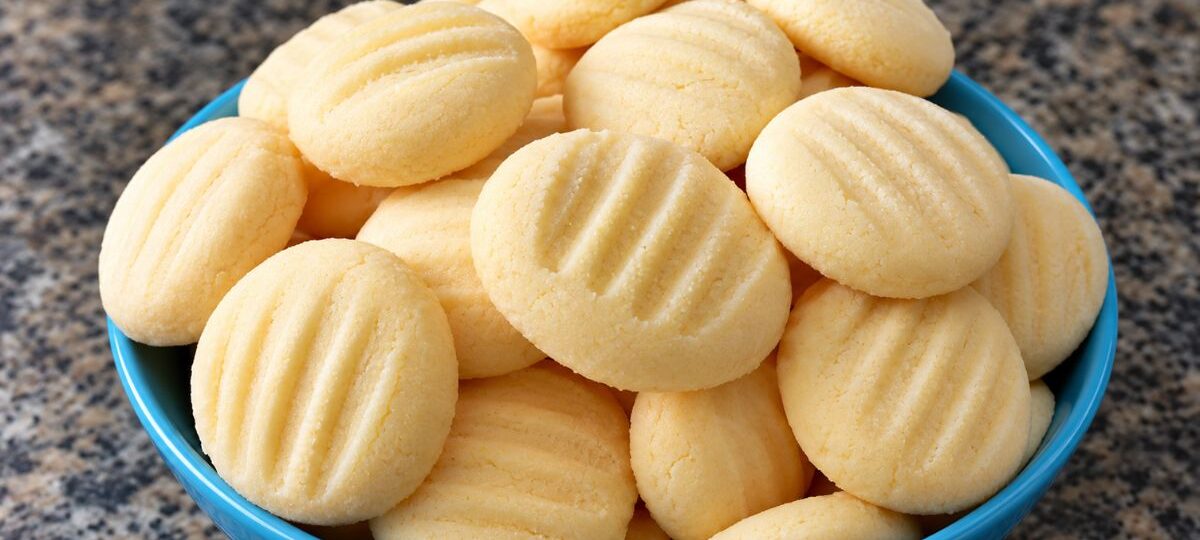 Uma tigela azul repleta de biscoitos de maizena artesanais redondos com marcas de garfo por cima apoiada em uma bancada de mármore escura.