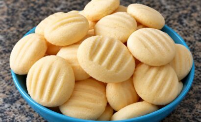 Uma tigela azul repleta de biscoitos de maizena artesanais redondos com marcas de garfo por cima apoiada em uma bancada de mármore escura.