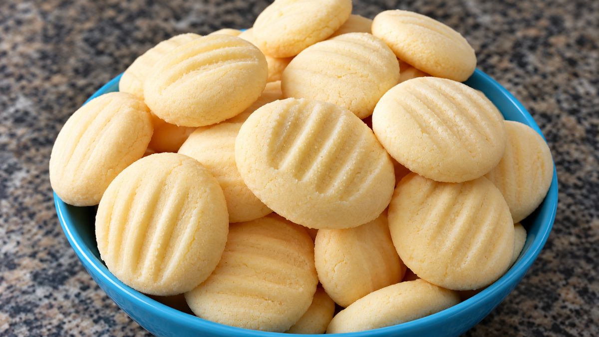 Uma tigela azul repleta de biscoitos de maizena artesanais redondos com marcas de garfo por cima apoiada em uma bancada de mármore escura.