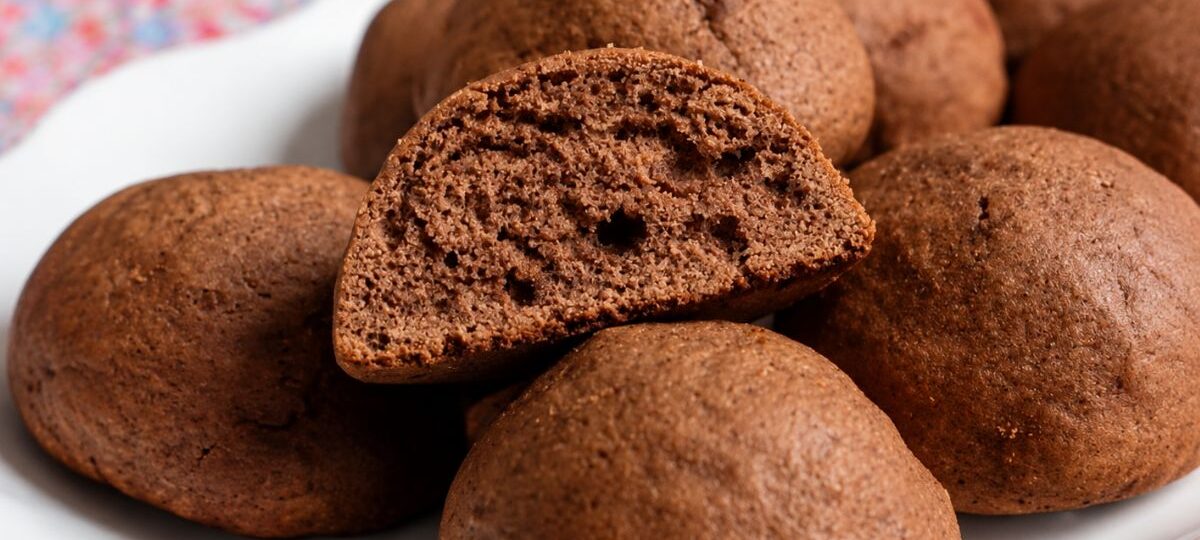 Um close em um prato branco cheio de biscoitos de melado escuros e arredondados. No centro, um biscoito está cortado ao meio revelando uma textura interna fofa e aerada. O fundo mostra uma toalha de mesa com estampa floral colorida borrada.