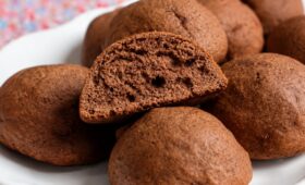Um close em um prato branco cheio de biscoitos de melado escuros e arredondados. No centro, um biscoito está cortado ao meio revelando uma textura interna fofa e aerada. O fundo mostra uma toalha de mesa com estampa floral colorida borrada.
