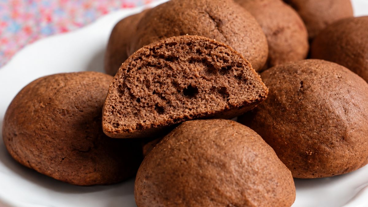 Um close em um prato branco cheio de biscoitos de melado escuros e arredondados. No centro, um biscoito está cortado ao meio revelando uma textura interna fofa e aerada. O fundo mostra uma toalha de mesa com estampa floral colorida borrada.