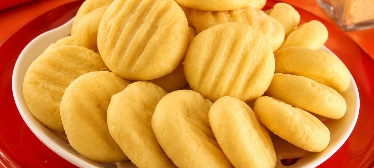 Biscoitos de queijo amarelos e arredondados com marcas de garfo dispostos em um prato branco sobre mesa vermelha.