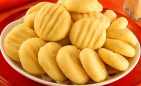 Biscoitos de queijo amarelos e arredondados com marcas de garfo dispostos em um prato branco sobre mesa vermelha.