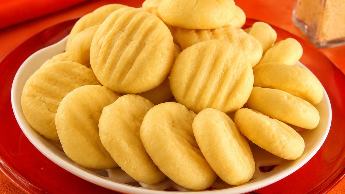 Biscoitos de queijo amarelos e arredondados com marcas de garfo dispostos em um prato branco sobre mesa vermelha.