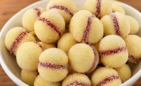 Tigela branca de cerâmica repleta de biscoitos casadinhos amanteigados recheados com goiabada vermelha e polvilhados com açúcar refinado, disposta sobre uma mesa de madeira.