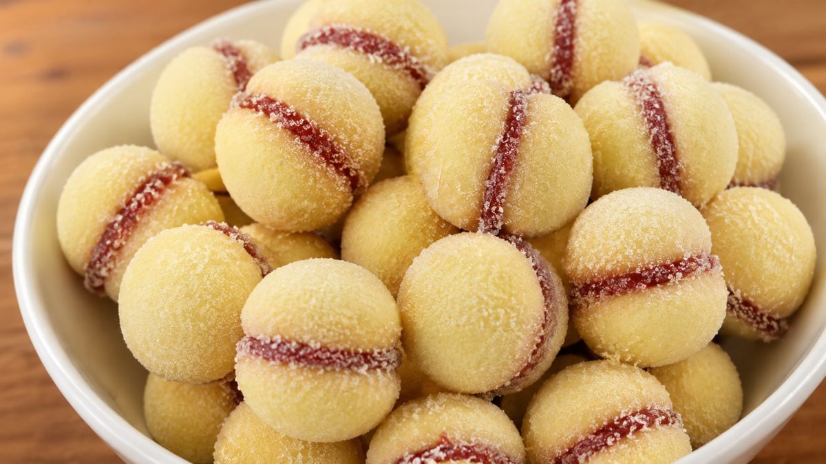 Tigela branca de cerâmica repleta de biscoitos casadinhos amanteigados recheados com goiabada vermelha e polvilhados com açúcar refinado, disposta sobre uma mesa de madeira.