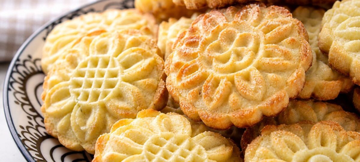 Close-up de uma pilha de biscoitos amanteigados caseiros com desenhos de flores douradas em um prato de cerâmica preta e branca.