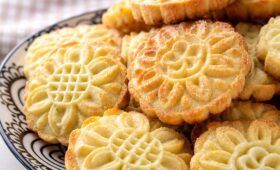 Close-up de uma pilha de biscoitos amanteigados caseiros com desenhos de flores douradas em um prato de cerâmica preta e branca.
