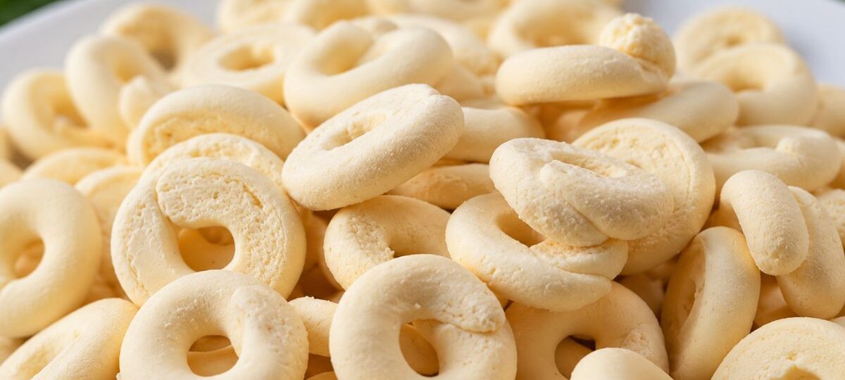 Um close em uma pilha de biscoitos de polvilho e leite condensado amarelos claros, em formato de rosca, em um prato branco.