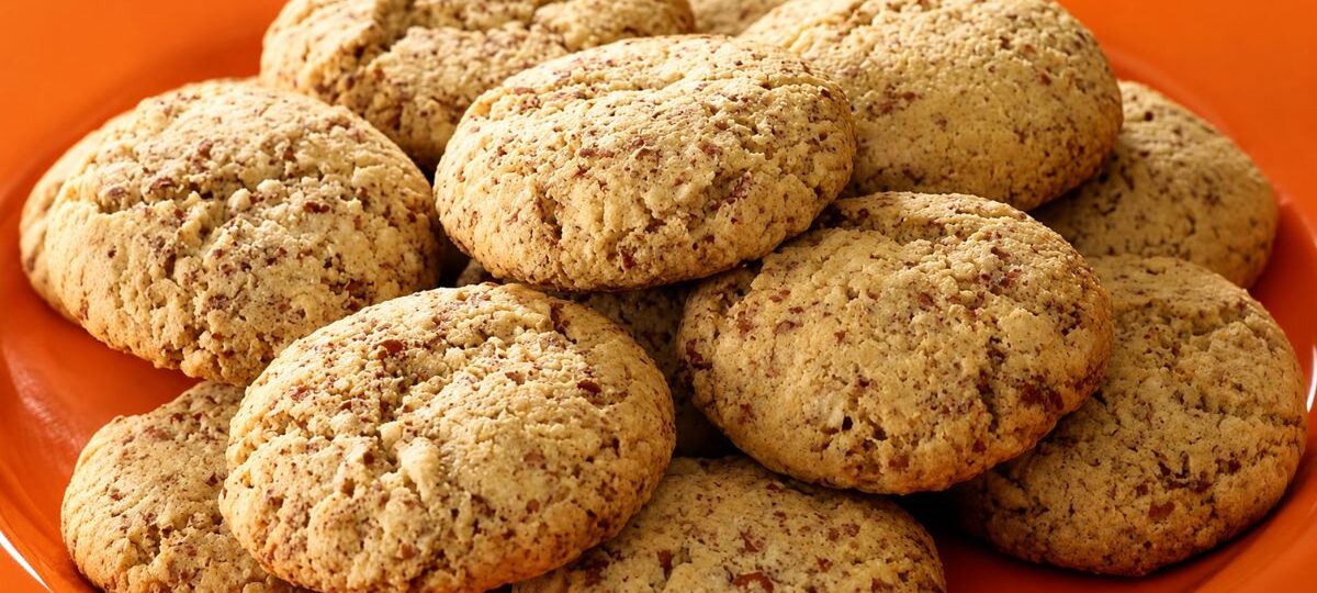 Uma vista de perto de uma pilha de biscoitos de amendoim macios e dourados, com pedaços visíveis de amendoim torrado, dispostos em um prato laranja vibrante.