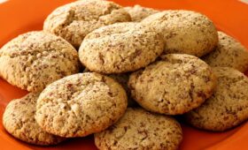 Uma vista de perto de uma pilha de biscoitos de amendoim macios e dourados, com pedaços visíveis de amendoim torrado, dispostos em um prato laranja vibrante.