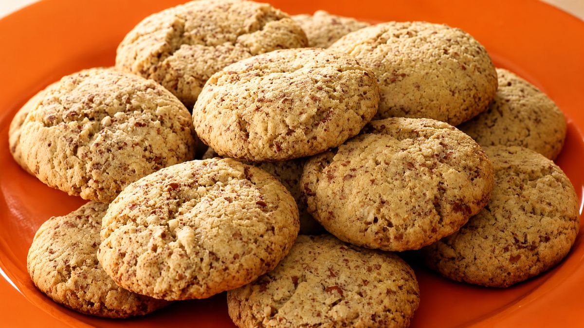 Uma vista de perto de uma pilha de biscoitos de amendoim macios e dourados, com pedaços visíveis de amendoim torrado, dispostos em um prato laranja vibrante.