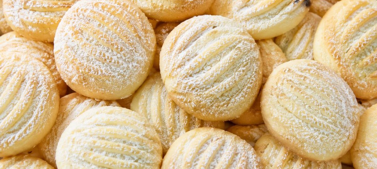 Close-up em plano superior de diversos biscoitos de baunilha redondos, com textura listrada e polvilhados com açúcar de confeiteiro, organizados em uma pilha.