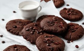 Uma pilha de biscoitos de chocolate caseiros redondos e achatados, cobertos com gotas de chocolate derretidas e pedaços rústicos de cacau cru, descansando sobre papel manteiga branco amassado em uma superfície de mármore claro. Ao fundo, uma pequena jarra de cerâmica branca contém leite fresco. Gotas de chocolate e cacau estão espalhadas pelo cenário.