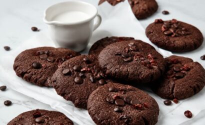 Uma pilha de biscoitos de chocolate caseiros redondos e achatados, cobertos com gotas de chocolate derretidas e pedaços rústicos de cacau cru, descansando sobre papel manteiga branco amassado em uma superfície de mármore claro. Ao fundo, uma pequena jarra de cerâmica branca contém leite fresco. Gotas de chocolate e cacau estão espalhadas pelo cenário.