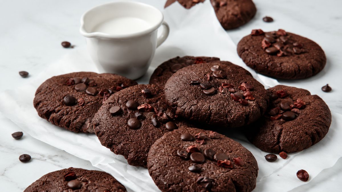Uma pilha de biscoitos de chocolate caseiros redondos e achatados, cobertos com gotas de chocolate derretidas e pedaços rústicos de cacau cru, descansando sobre papel manteiga branco amassado em uma superfície de mármore claro. Ao fundo, uma pequena jarra de cerâmica branca contém leite fresco. Gotas de chocolate e cacau estão espalhadas pelo cenário.