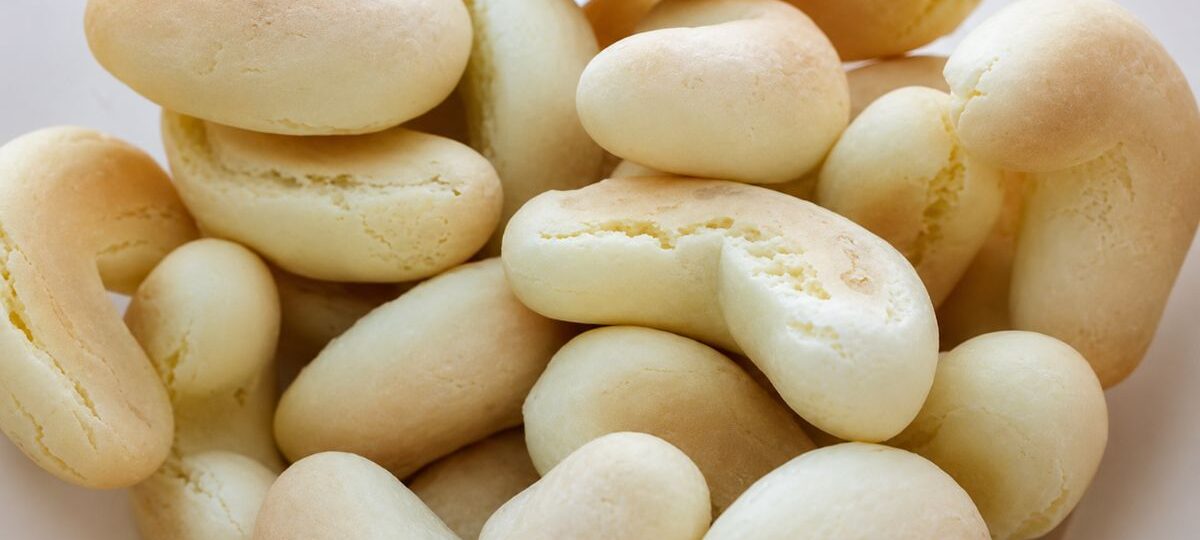 Uma foto de plano superior em close-up de um aglomerado de biscoitos de polvilho doces em formato de ferradura em uma tigela branca rústica. Os biscoitos são de cor bege claro, ligeiramente texturizados com algumas rachaduras superficiais e são amontoados, criando um padrão dinâmico de formas curvas e alongadas.