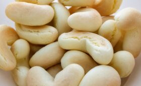 Uma foto de plano superior em close-up de um aglomerado de biscoitos de polvilho doces em formato de ferradura em uma tigela branca rústica. Os biscoitos são de cor bege claro, ligeiramente texturizados com algumas rachaduras superficiais e são amontoados, criando um padrão dinâmico de formas curvas e alongadas.