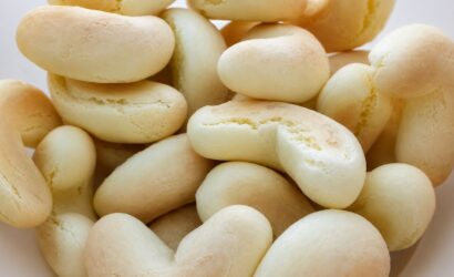 Uma foto de plano superior em close-up de um aglomerado de biscoitos de polvilho doces em formato de ferradura em uma tigela branca rústica. Os biscoitos são de cor bege claro, ligeiramente texturizados com algumas rachaduras superficiais e são amontoados, criando um padrão dinâmico de formas curvas e alongadas.
