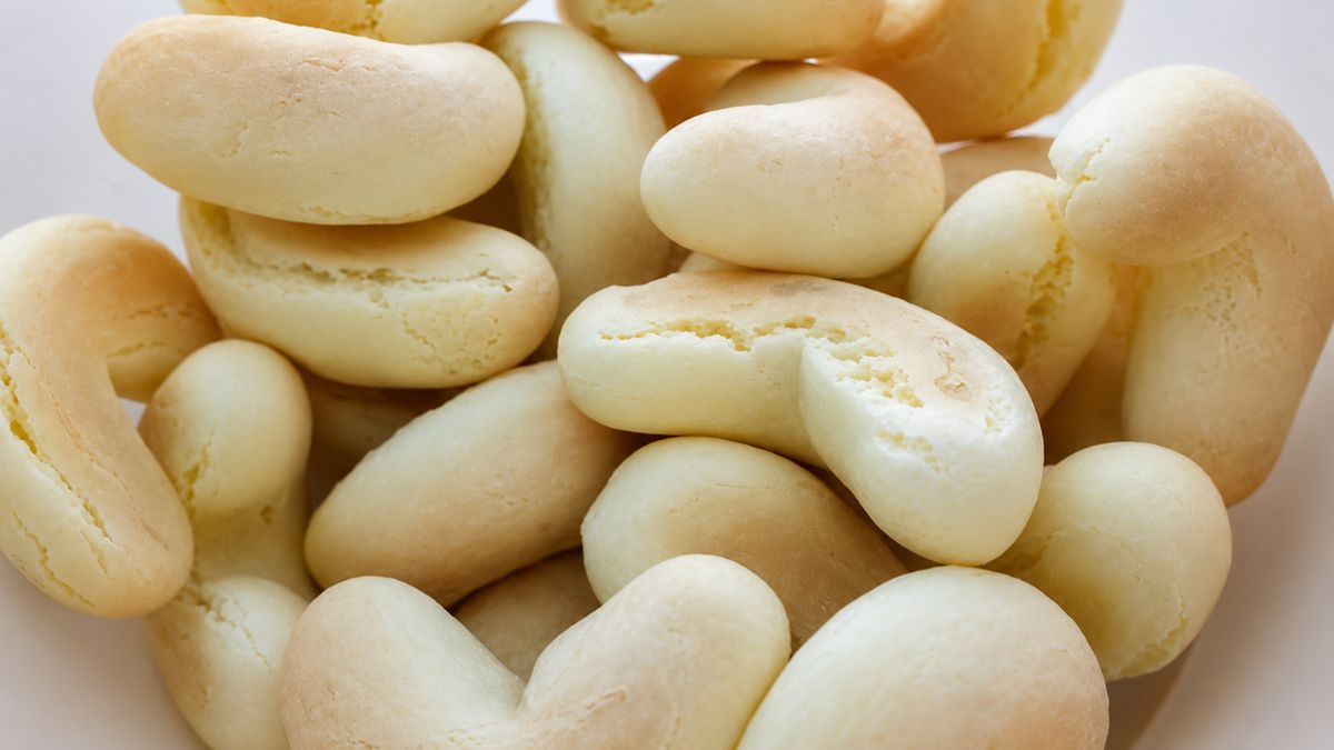 Uma foto de plano superior em close-up de um aglomerado de biscoitos de polvilho doces em formato de ferradura em uma tigela branca rústica. Os biscoitos são de cor bege claro, ligeiramente texturizados com algumas rachaduras superficiais e são amontoados, criando um padrão dinâmico de formas curvas e alongadas.