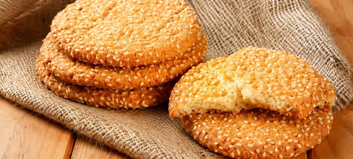 Quatro biscoitos redondos artesanais cobertos com gergelim em um pano de estopa rústico sobre uma mesa de madeira. Um biscoito está partido ao meio, revelando a textura interior.