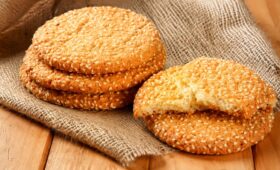 Quatro biscoitos redondos artesanais cobertos com gergelim em um pano de estopa rústico sobre uma mesa de madeira. Um biscoito está partido ao meio, revelando a textura interior.