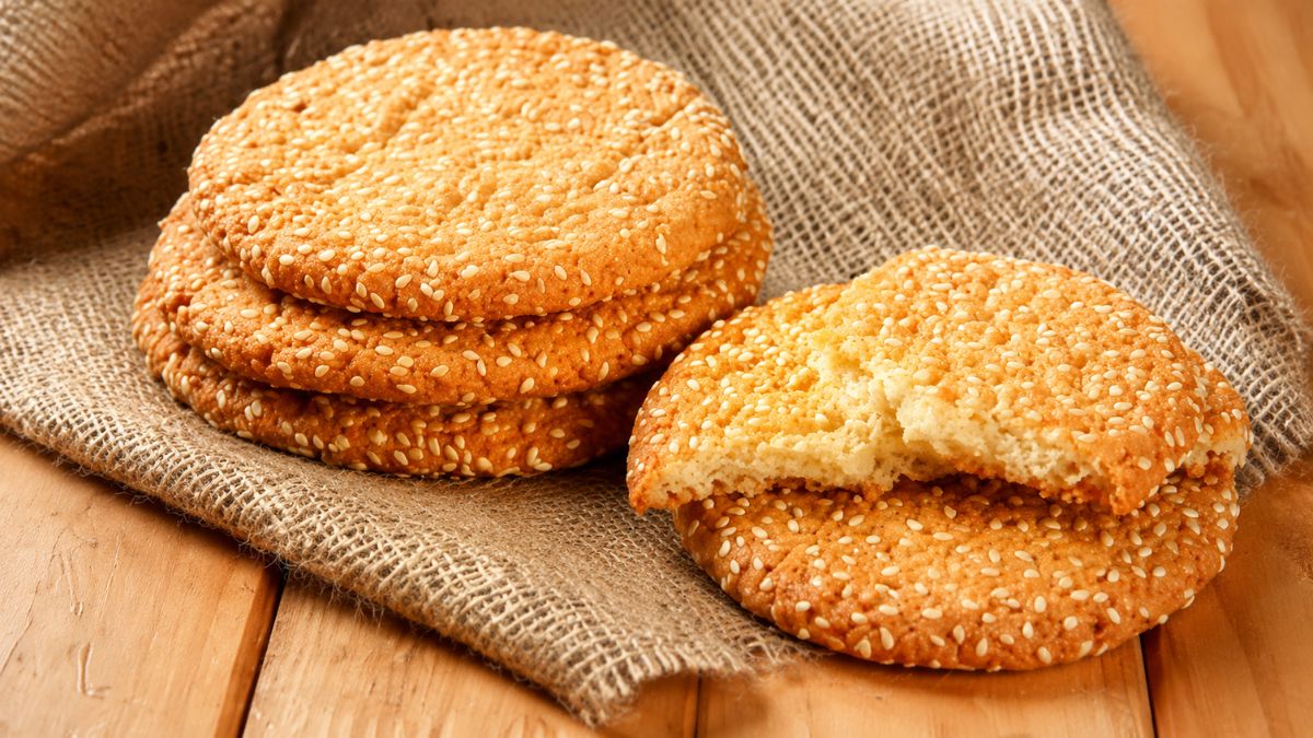 Quatro biscoitos redondos artesanais cobertos com gergelim em um pano de estopa rústico sobre uma mesa de madeira. Um biscoito está partido ao meio, revelando a textura interior.